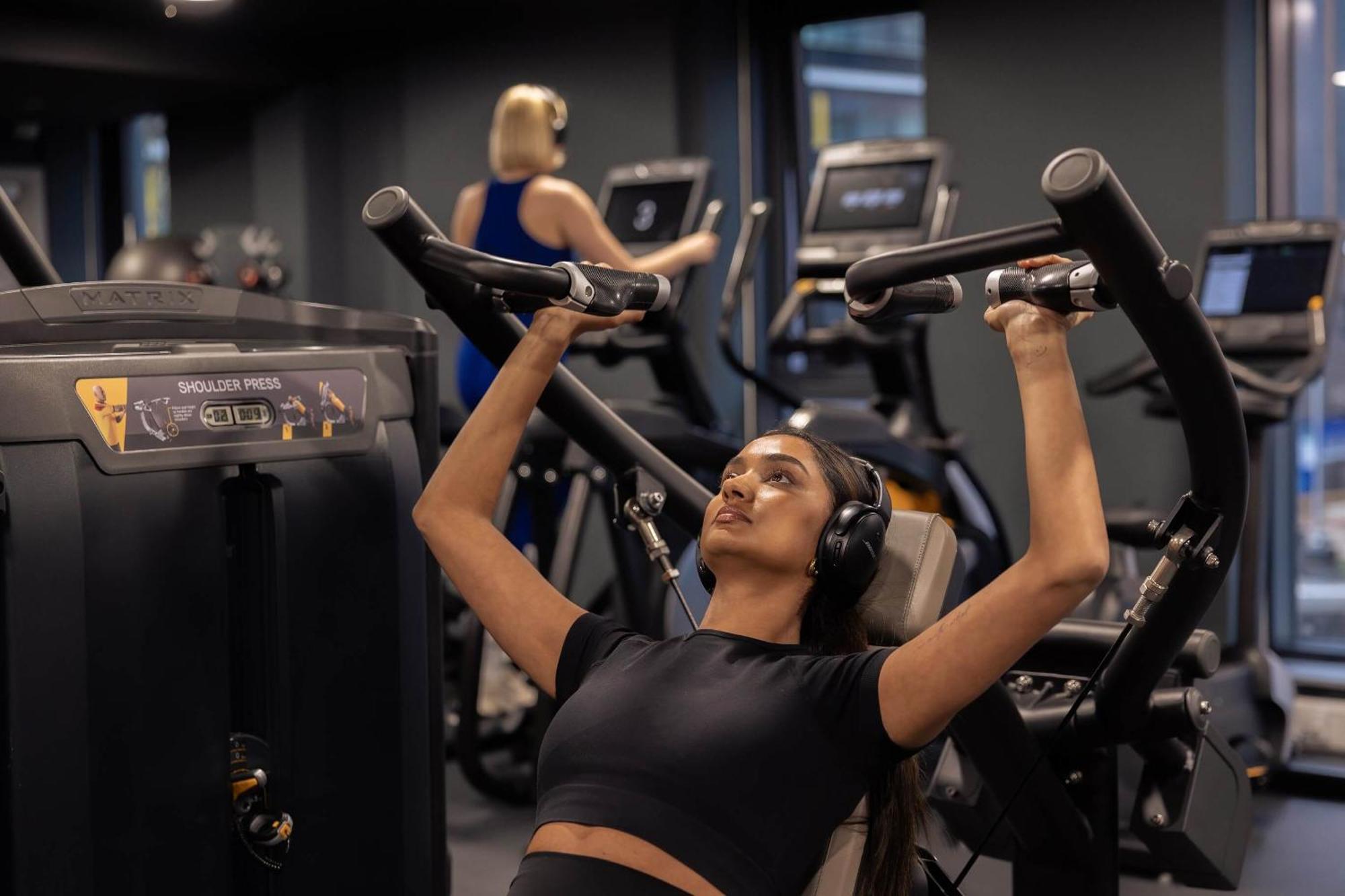 Citysuites 2 Aparthotel Salford (Greater Manchester) Exterior foto The photo shows a gym setting with two individuals engaged in physical exercise. In the foreground, a woman is using a chest press machine while wearing headphones, focusing on her workout. She appears to be in a seated position, pushing the handles 