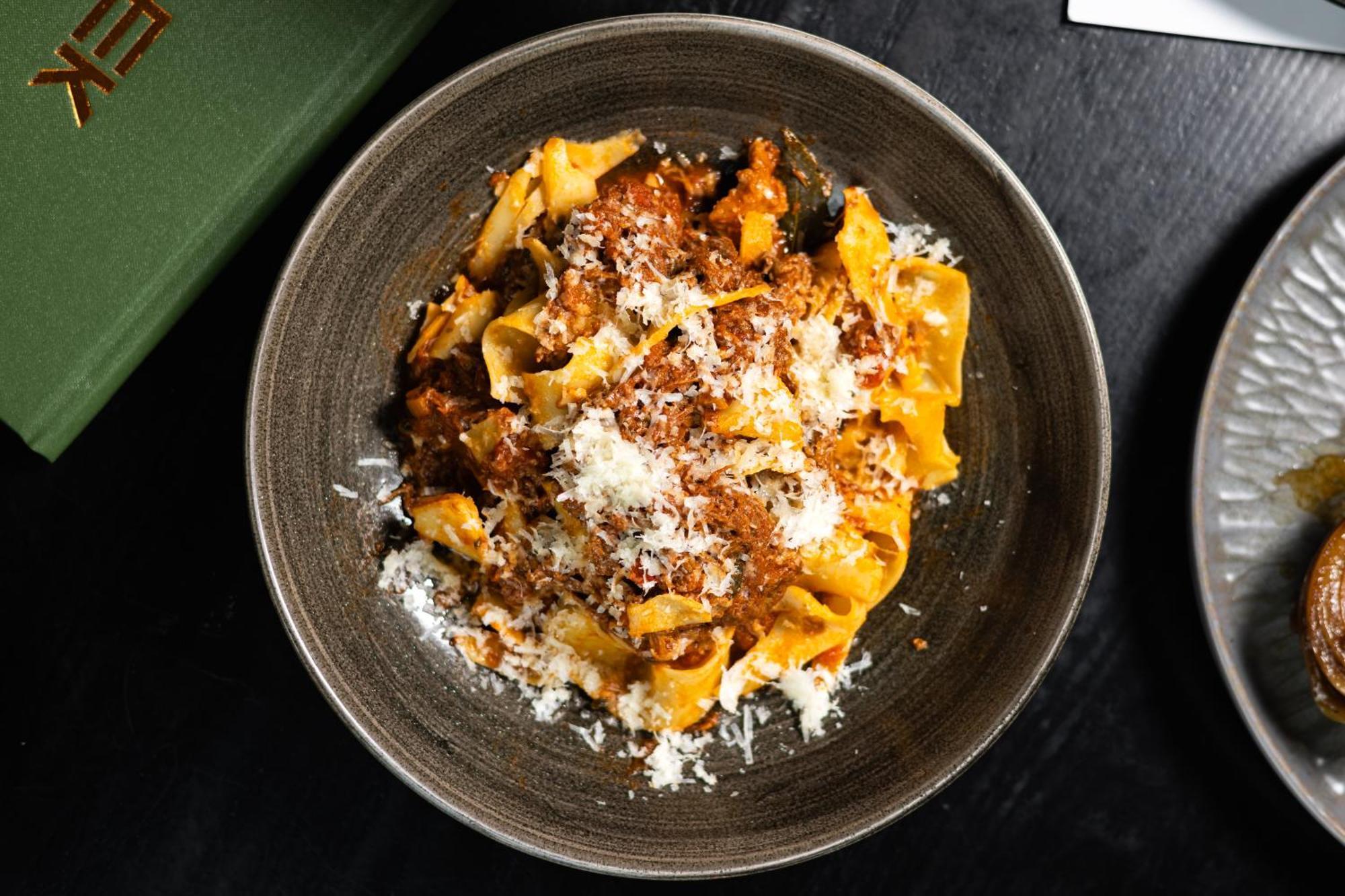 Citysuites 2 Aparthotel Salford (Greater Manchester) Exterior foto The photo shows a bowl of pasta featuring wide, ribbon-like noodles, likely pappardelle, fully coated in a rich meat sauce. The dish is garnished with a generous sprinkling of grated cheese, adding a creamy texture to the presentation. The dark bowl 